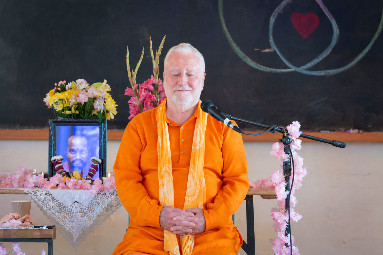 Swami Brahmananda seated in orange robes with his hands together beside flowers and a microphone