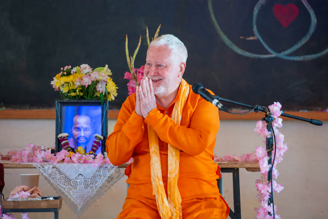 Swami Brahmananda greeting people with his hands together, seated in orange robes beside flowers and a microphone